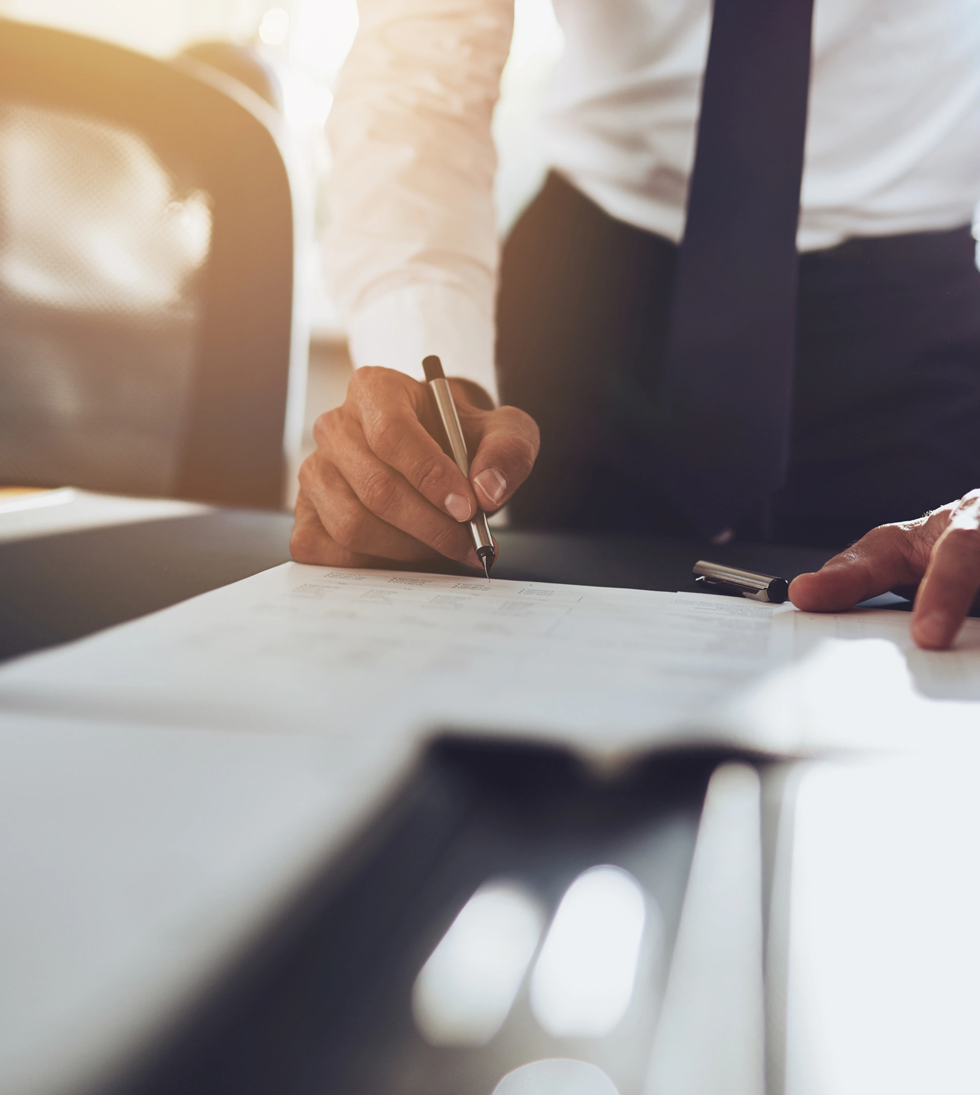 A person in business attire leans over a desk, holding a pen and writing on a document, with sunlight streaming in from the background.