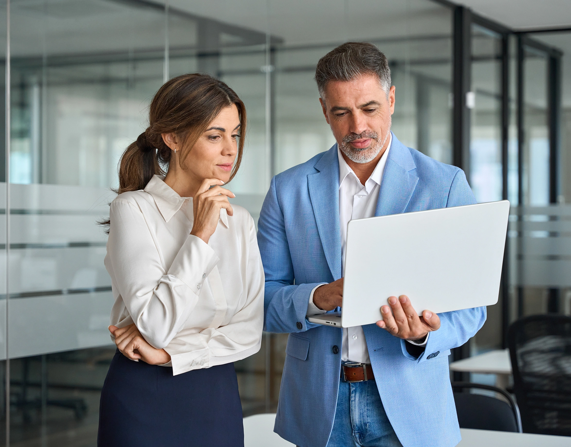 A woman and a man stand in a modern office, looking intently at a laptop screen. The man holds the laptop while the woman stands beside him, appearing thoughtful-mobile
