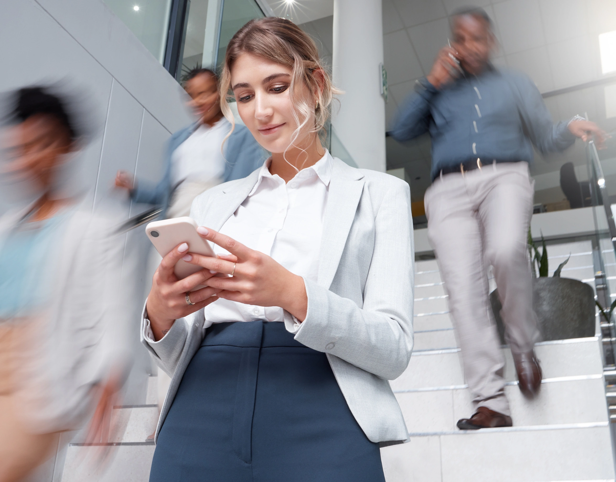 A woman in business attire stands on stairs looking at her smartphone, while several people walk past her in motion blur-moble
