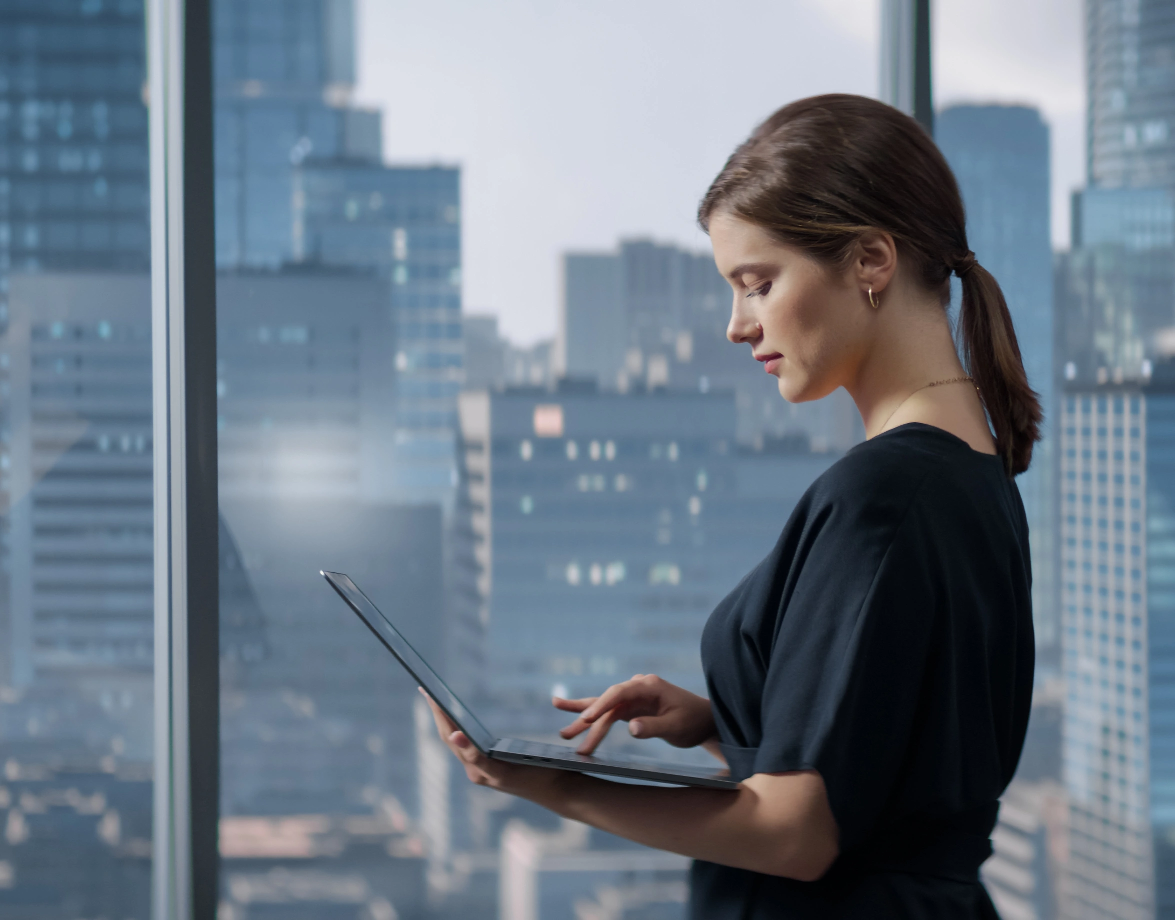 A woman stands by a large window in an office, holding and using a laptop. Tall city buildings are visible outside the window in the background-mobile