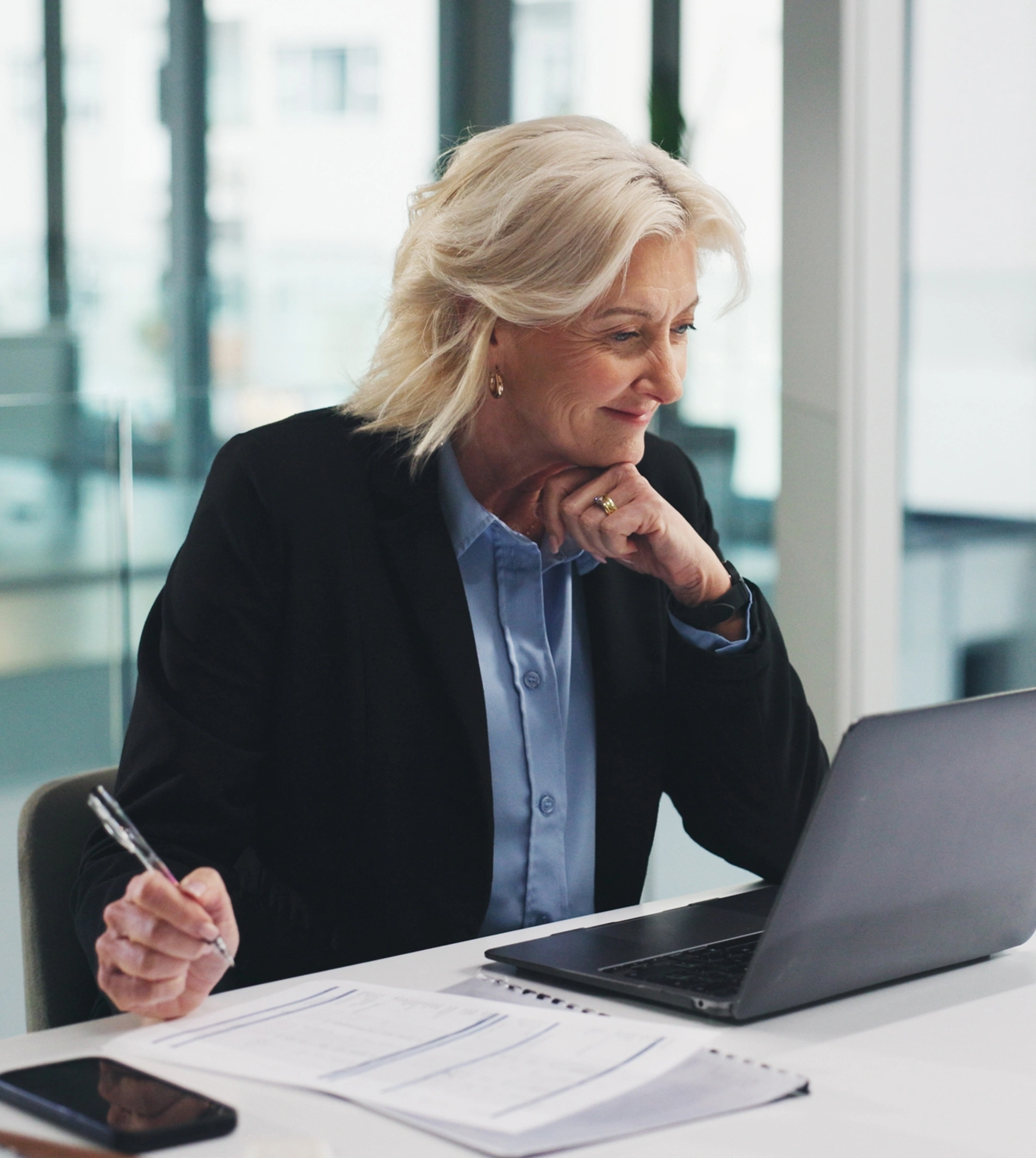 An older woman in a black blazer sits at a desk, looking at a laptop and holding a pen. She appears focused and engaged in her work in a modern office setting.