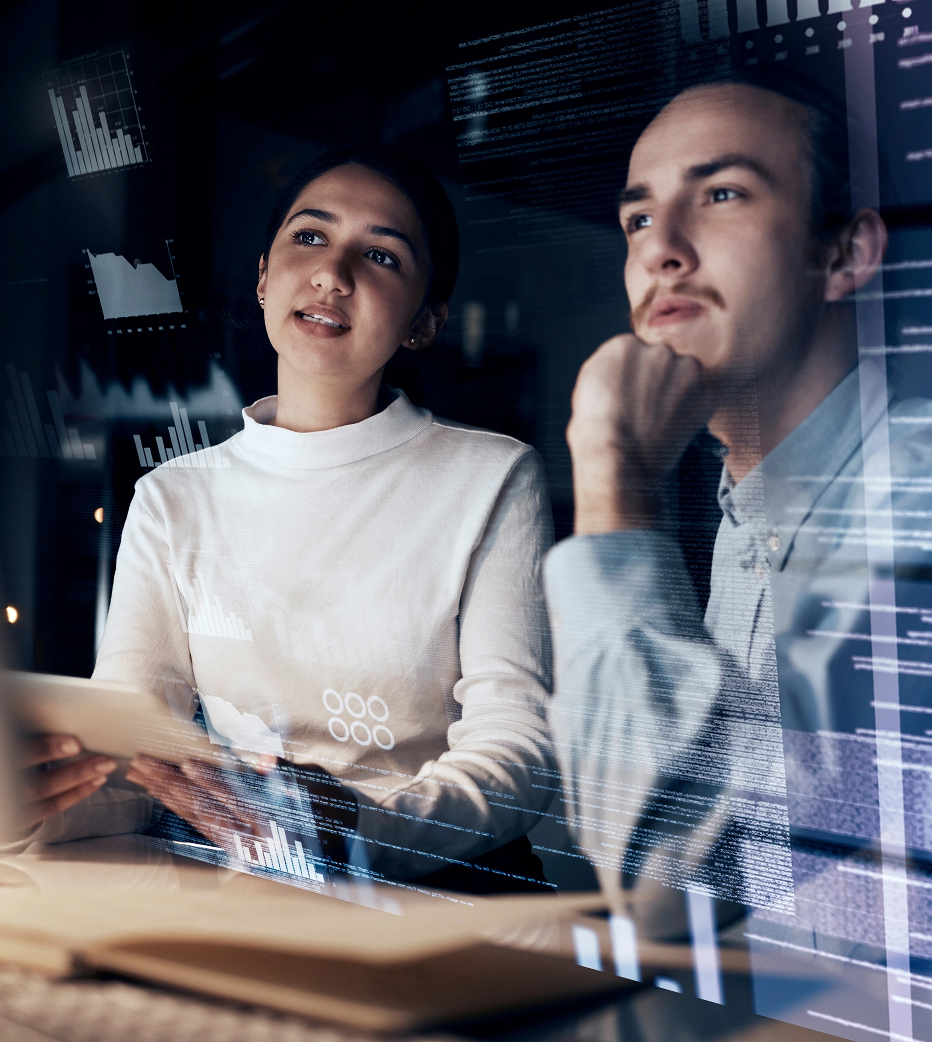 Two people sit together, looking thoughtfully at data charts and graphs projected on a transparent screen in a dimly lit room.