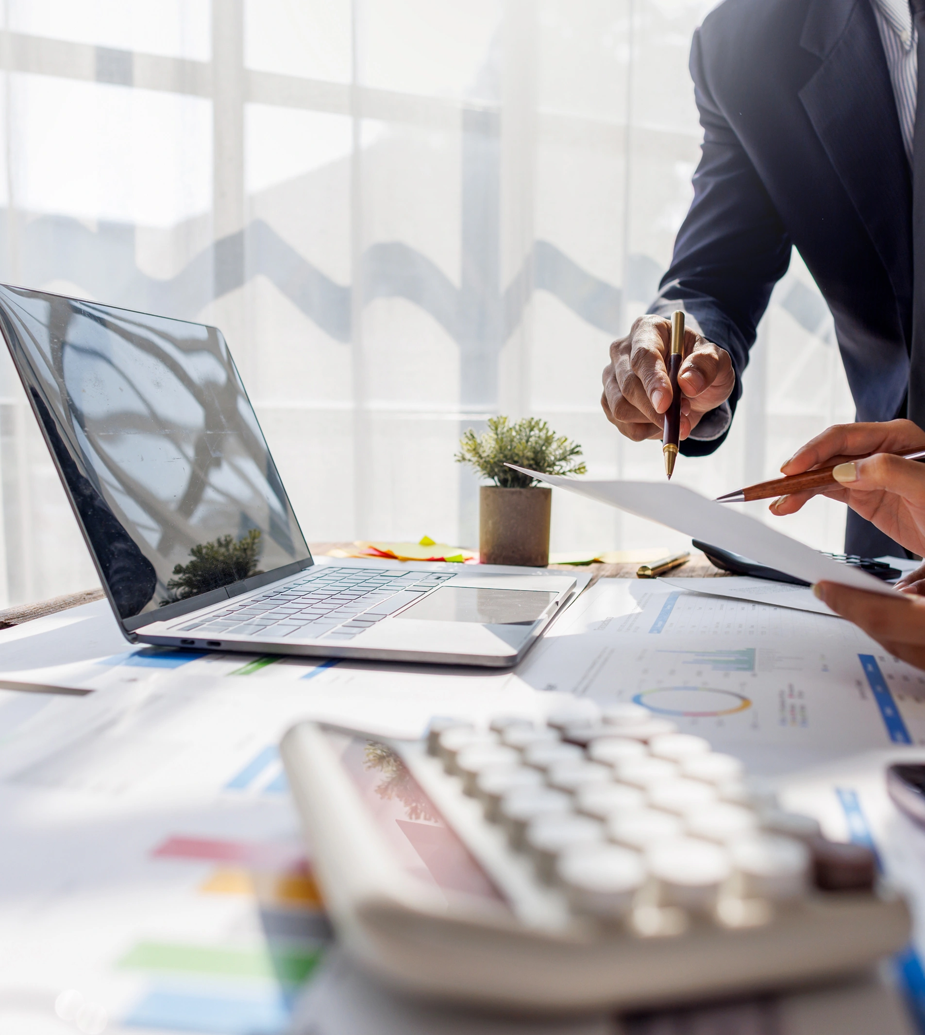 A close-up view of a desk with a laptop, calculator, and financial documents, where two people in professional attire are reviewing papers and discussing them with pens.