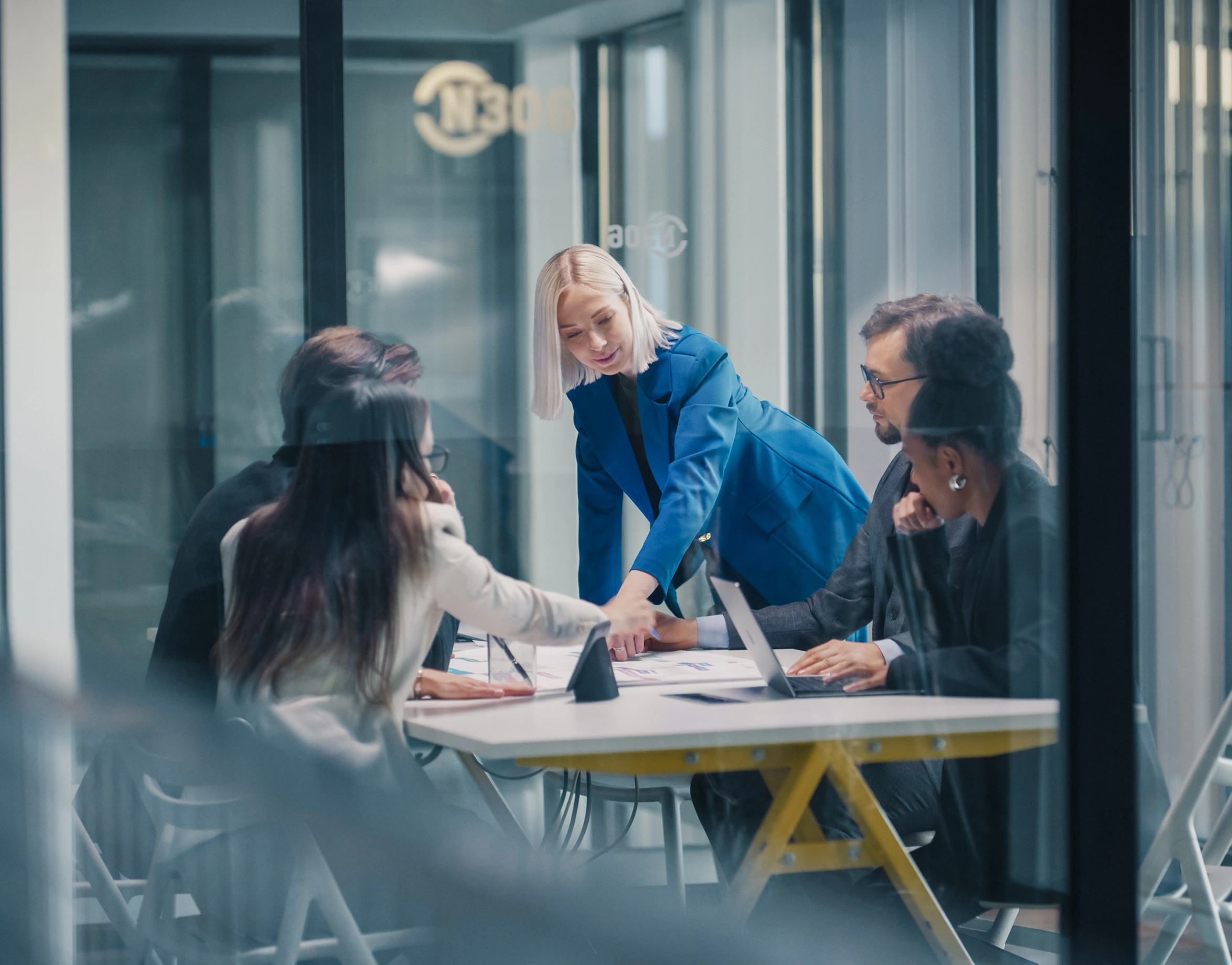 A diverse group of business professionals collaborate around a table in a modern office, with one person in a blue blazer gesturing towards documents.