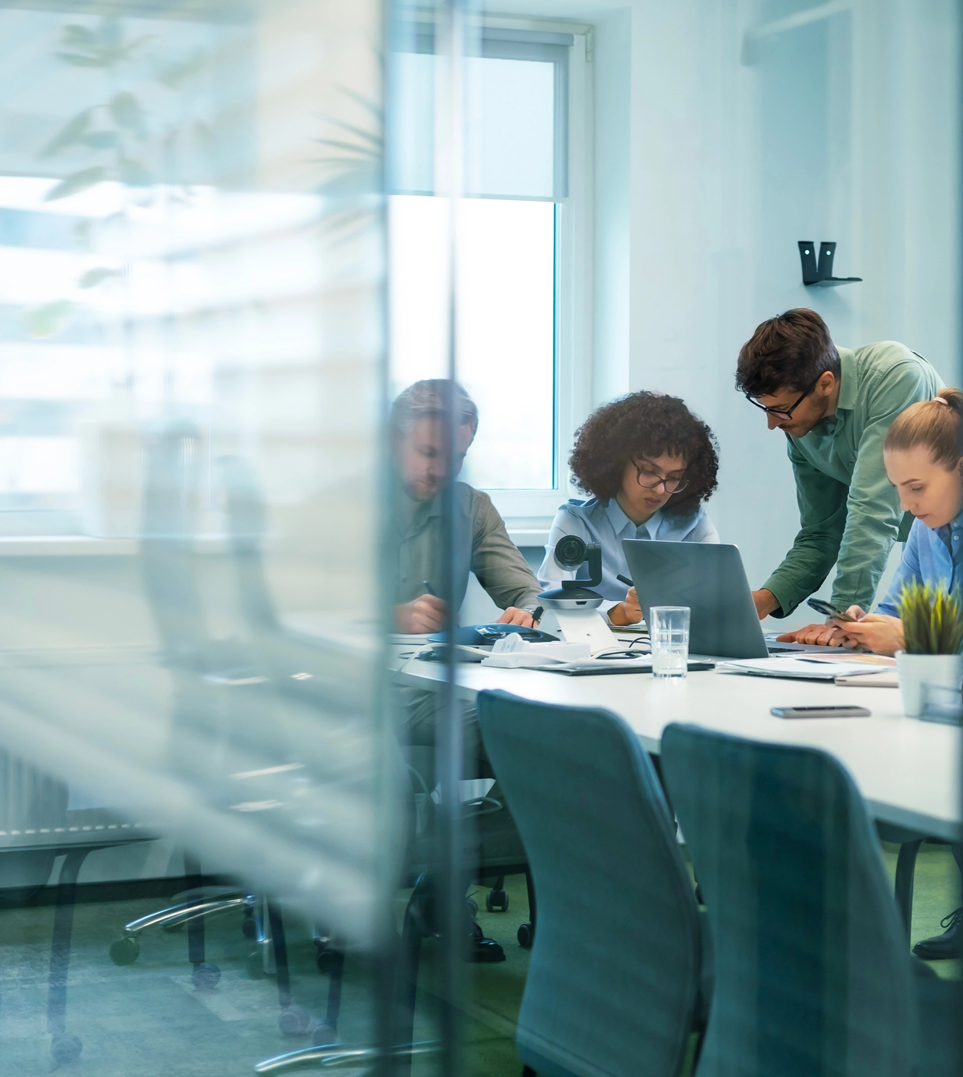A diverse group of colleagues collaborates around a conference table, with one person leaning in to observe a laptop.