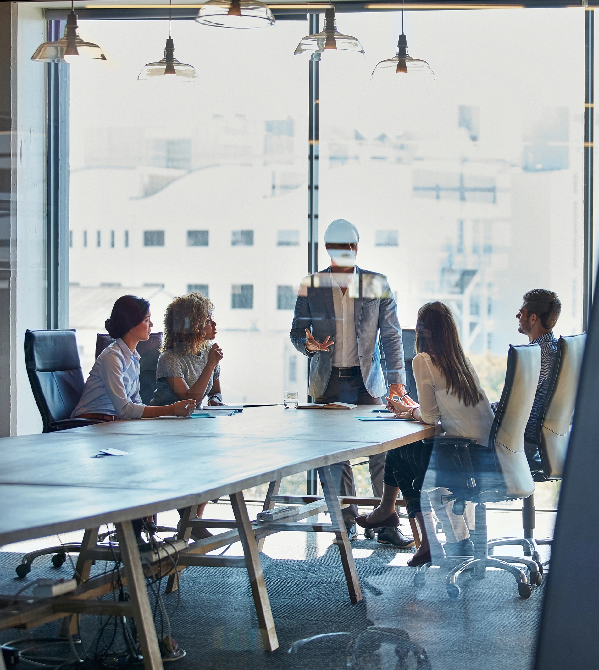 A diverse group of professionals participates in a business meeting around a large table in a modern conference room, with a presenter standing and gesturing while facing attendees.