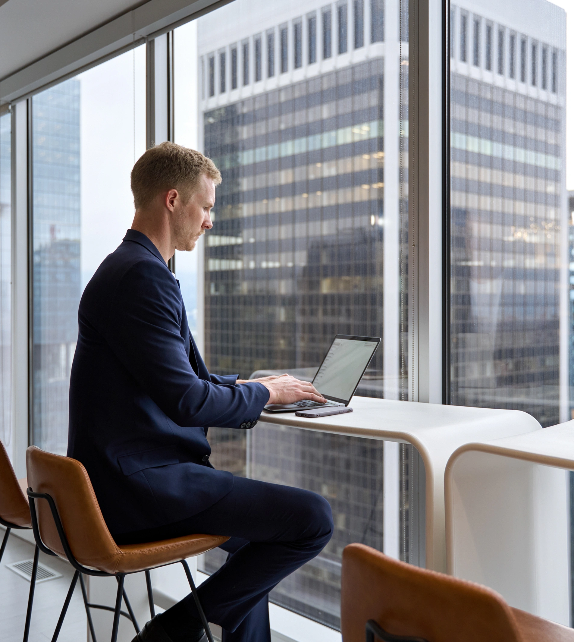 A man in a suit works on a laptop at a high table by a window overlooking tall office buildings in a city.