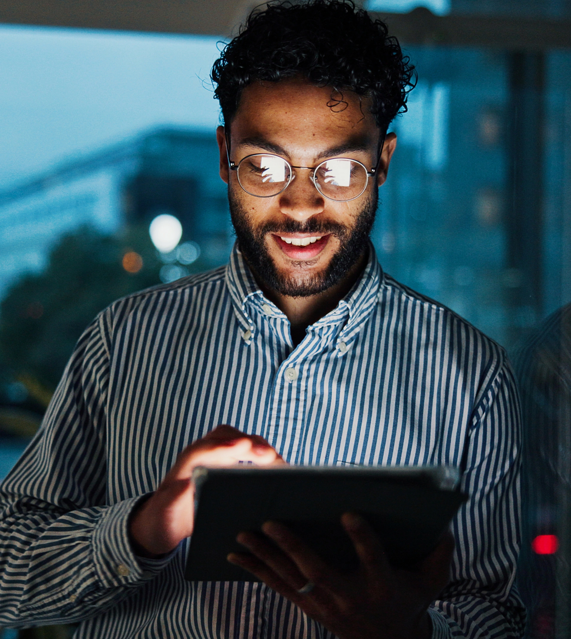 A man with curly hair and glasses smiles as he looks at a tablet in front of a window with blurred city lights at night.