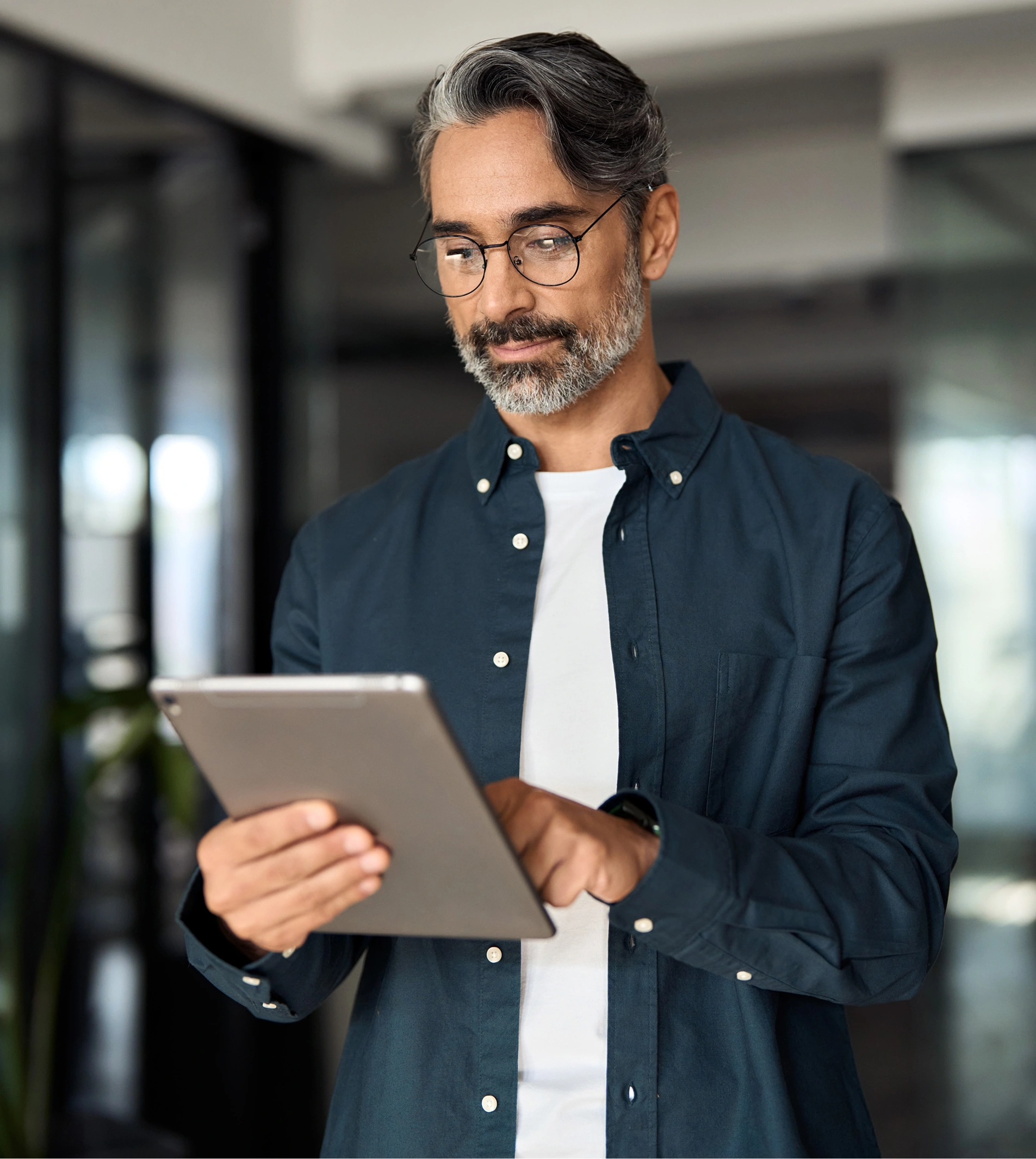 A man with graying hair and a beard, wearing glasses and a dark blue collared shirt over a white t-shirt, holds and looks at a tablet device in his hands.