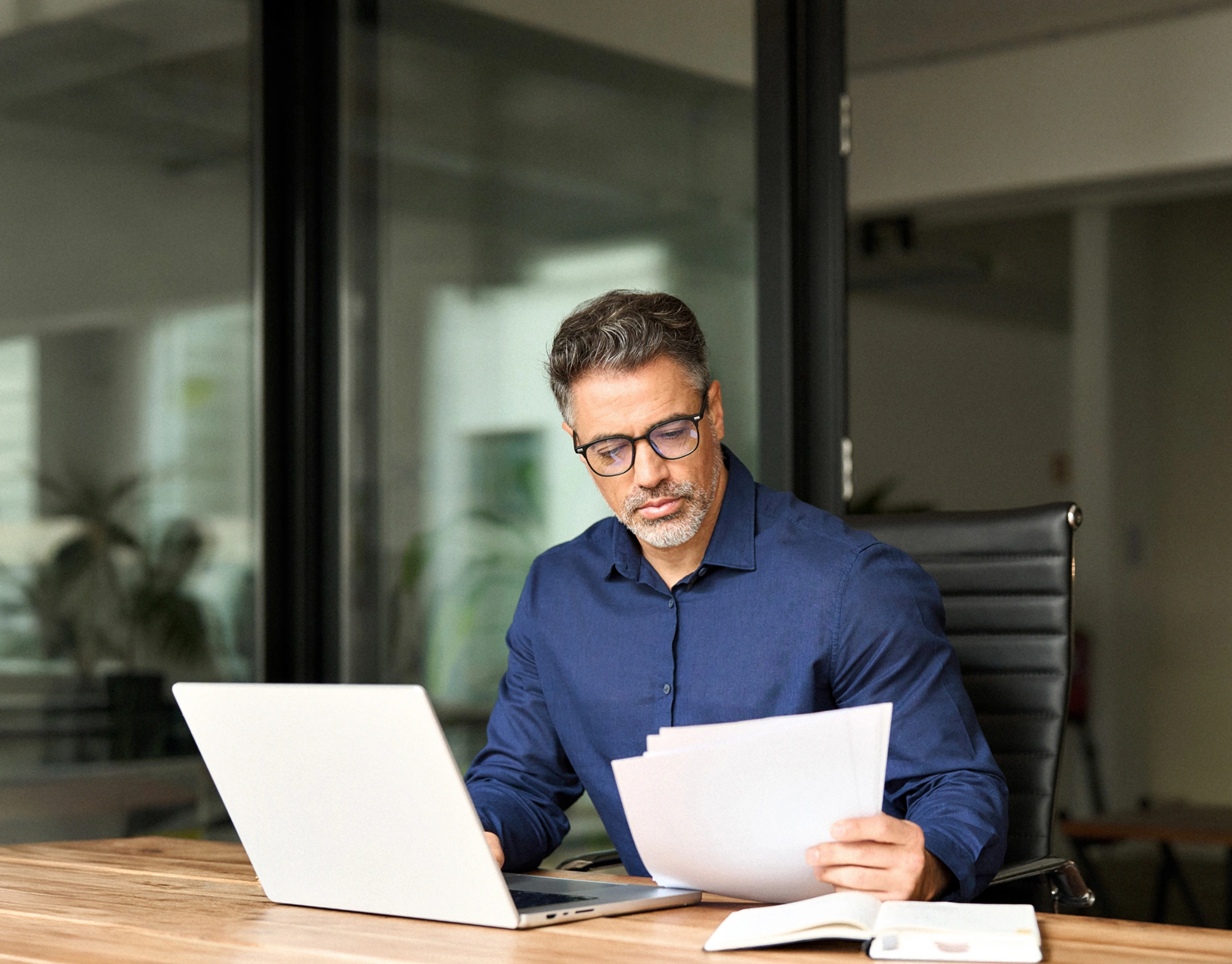A middle-aged man with glasses and salt-and-pepper hair, wearing a blue dress shirt, sits at a wooden desk in an office. He is looking down at papers in his hands while a laptop is open in front