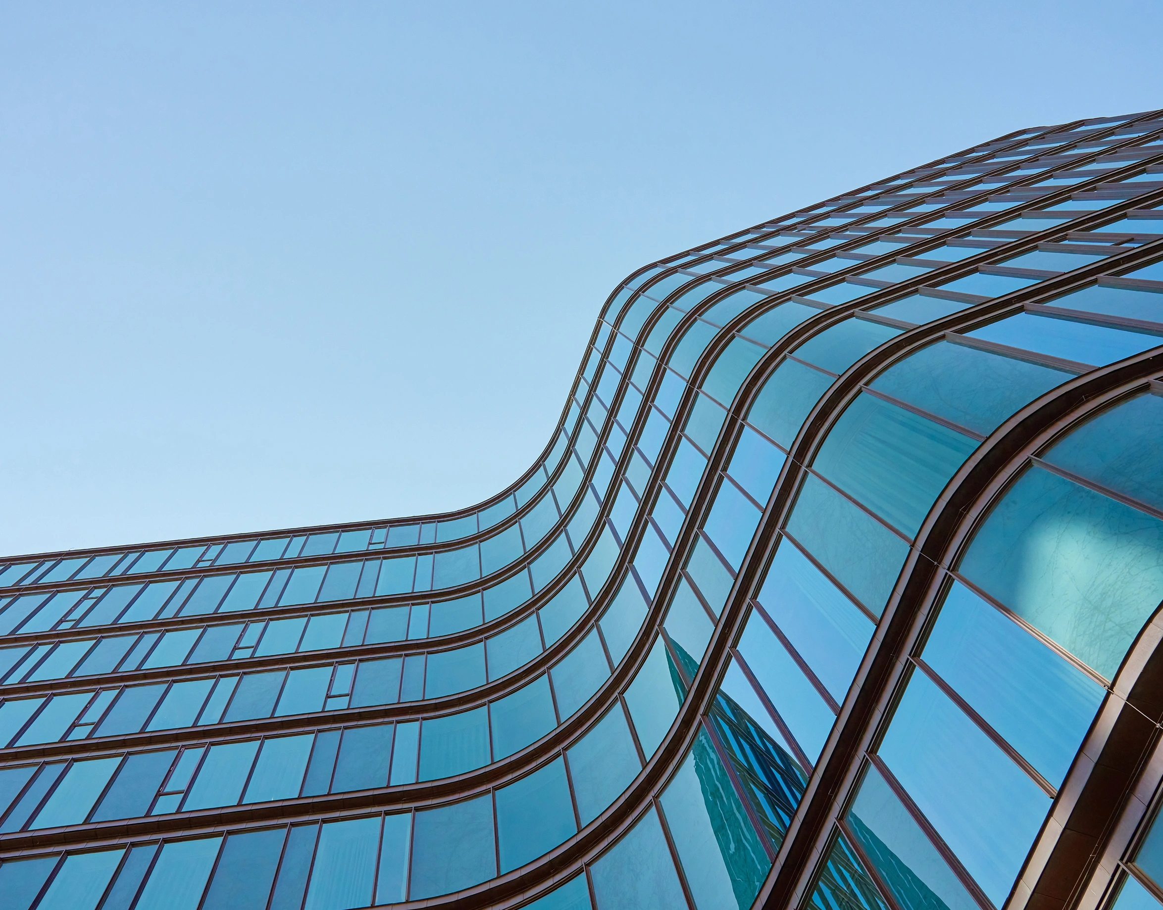 A modern building facade with a wavy, organic design made of reflective blue glass panels and dark framing against a clear sky.