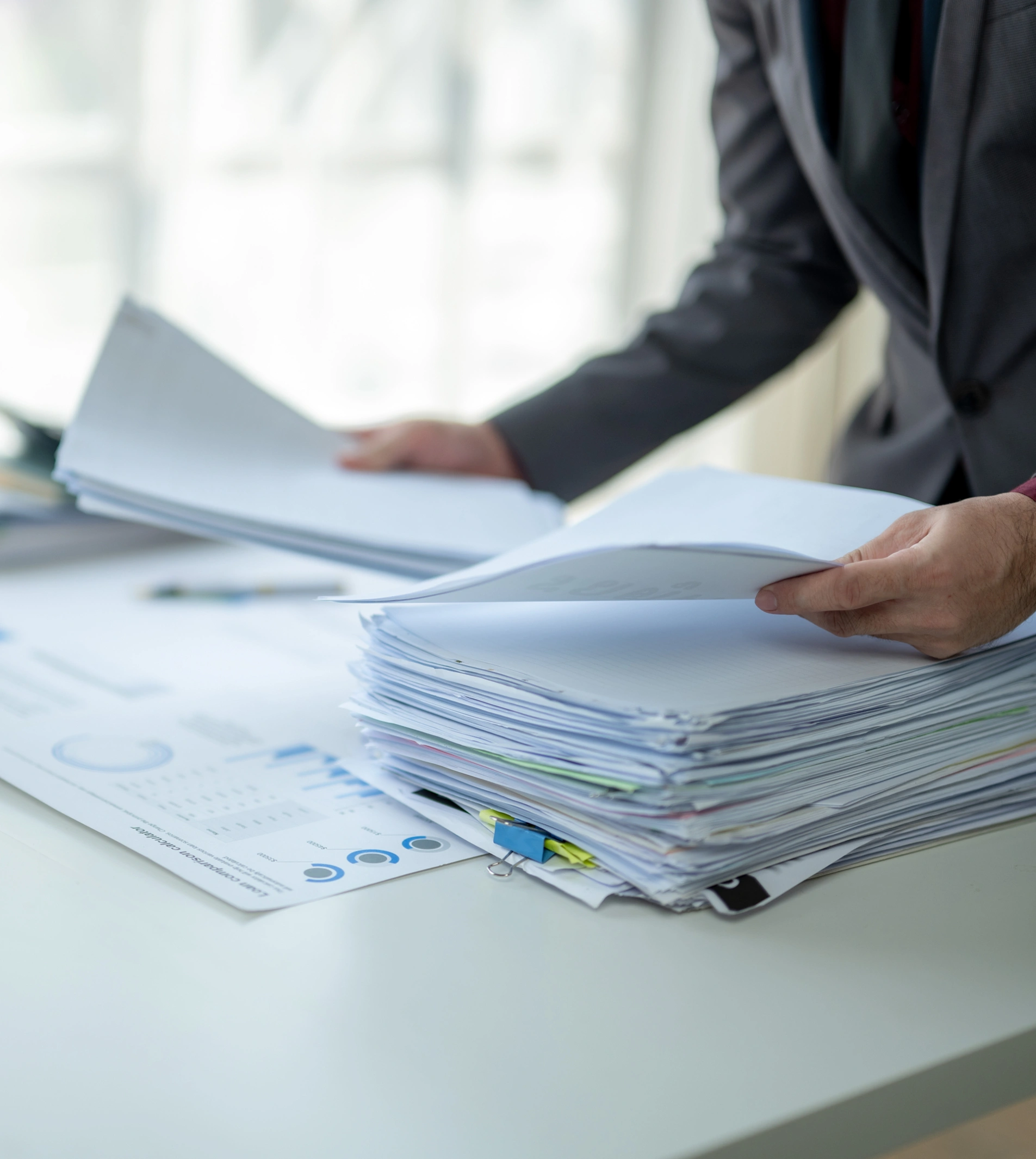 A person in a dark suit is reviewing a large stack of documents on a white desk, with charts and graphs visible on some pages.