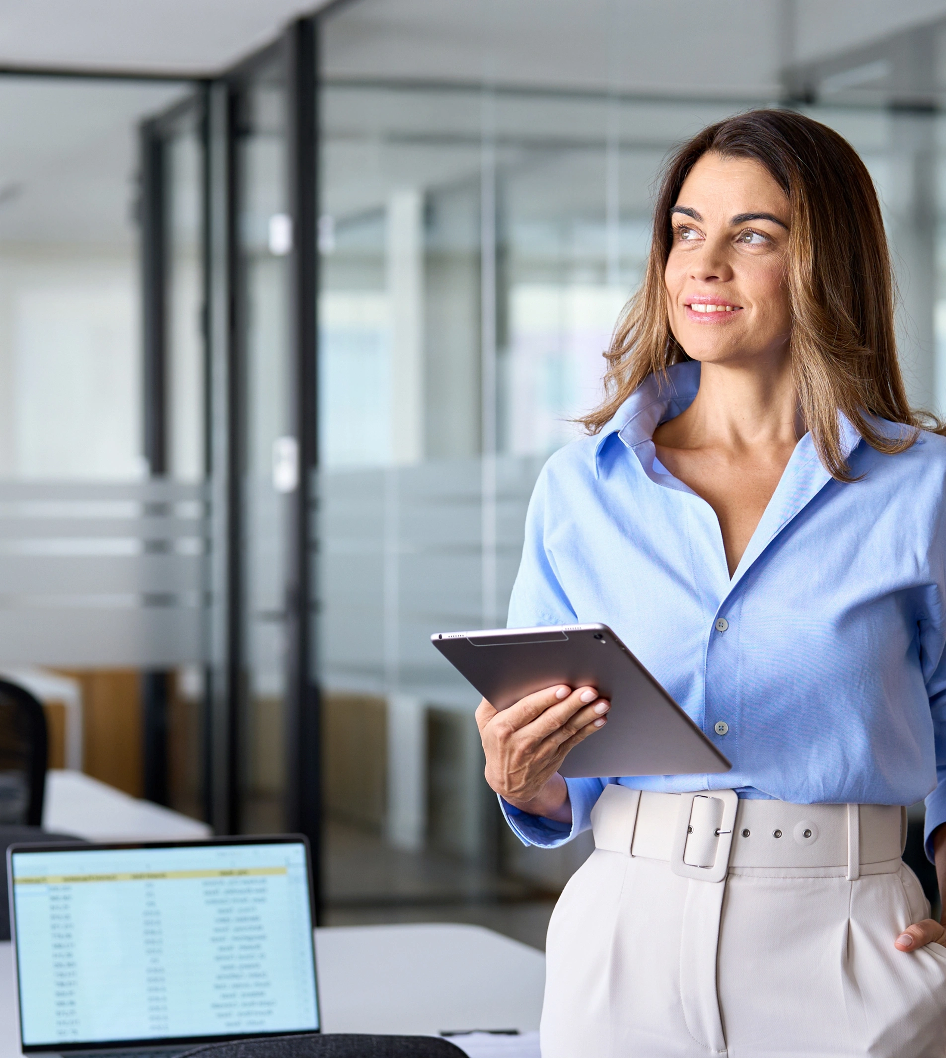 A professional woman in a blue button-down shirt and white trousers holds a tablet while looking to the side in an office setting.