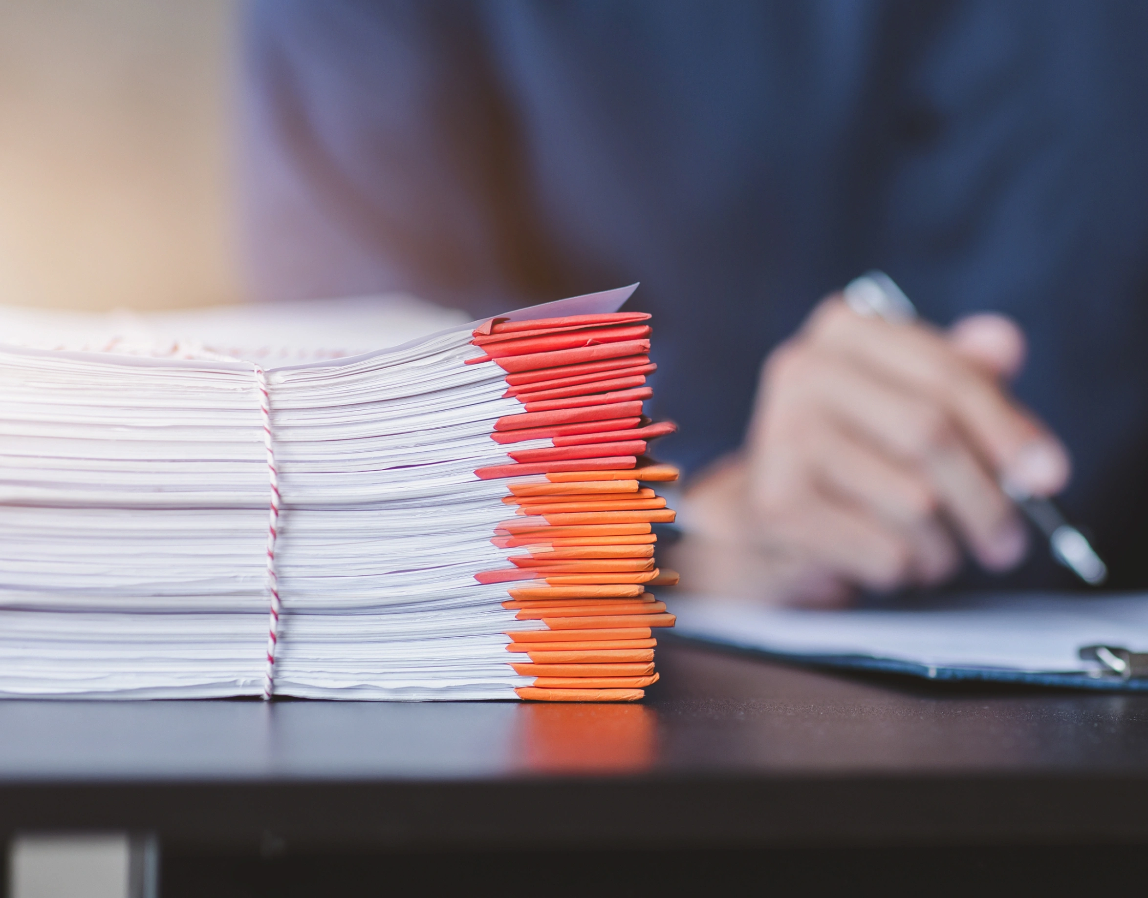 A tall stack of documents tied with twine, with red and orange folders visible, sits on a desk in front of a person holding a pen to a clipboard.