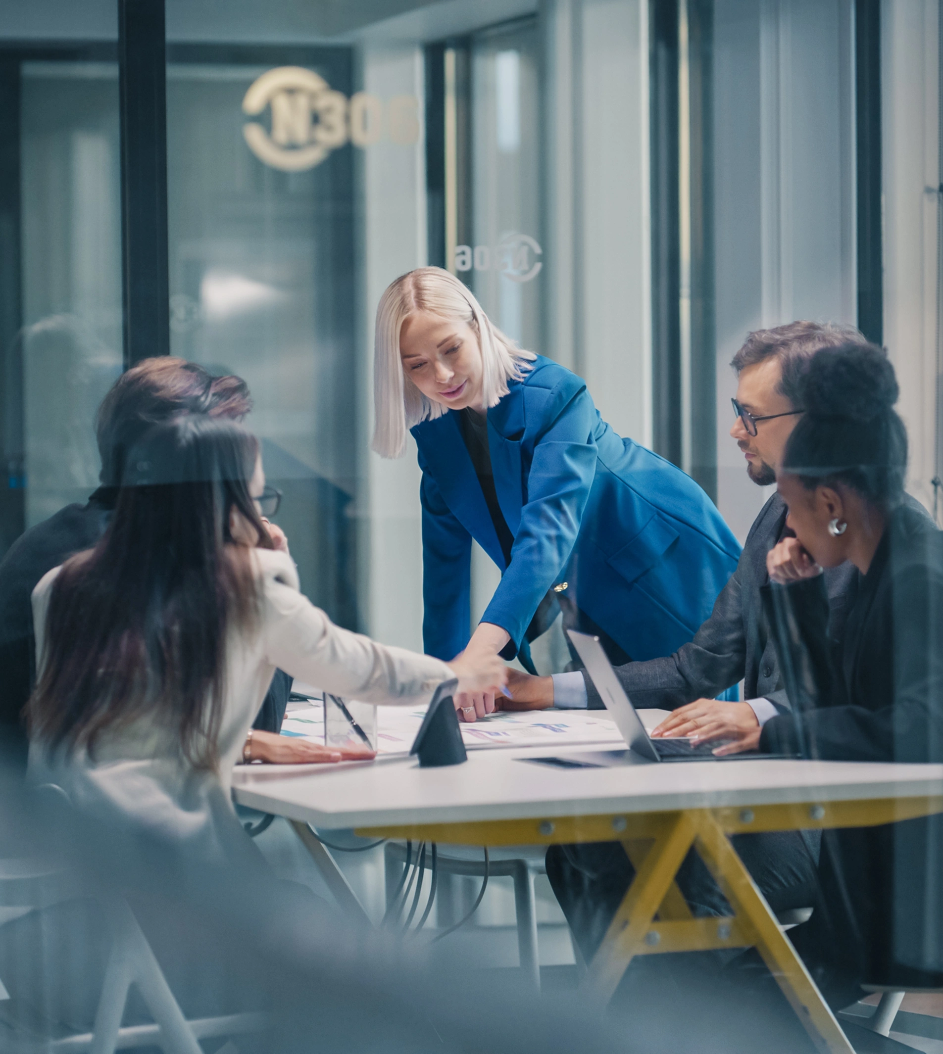 A woman in a blue blazer leads a meeting with four colleagues around a table in a modern office conference room.