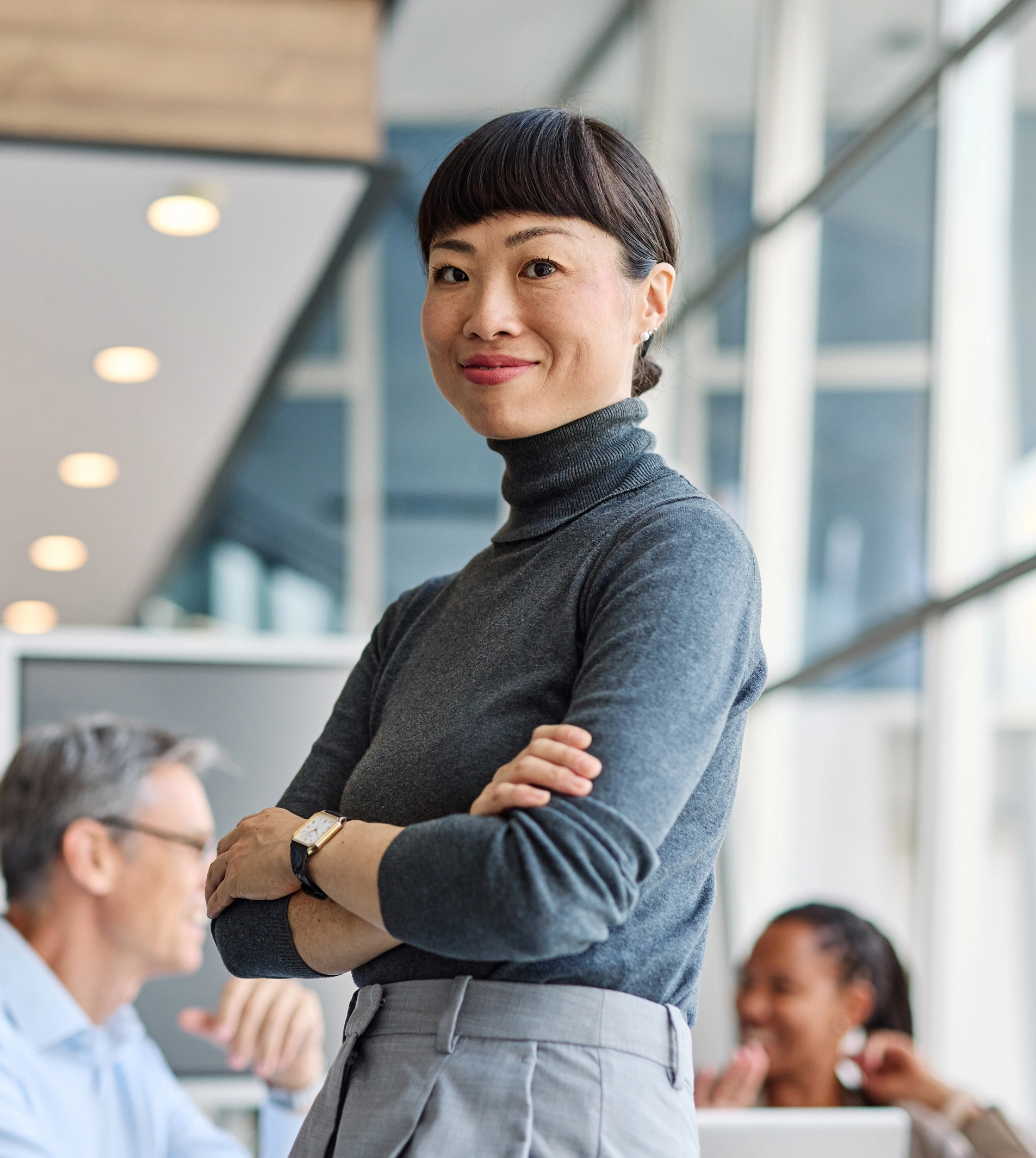 An Asian woman wearing a grey turtleneck and grey pants stands with her arms crossed in an office setting, smiling confidently.