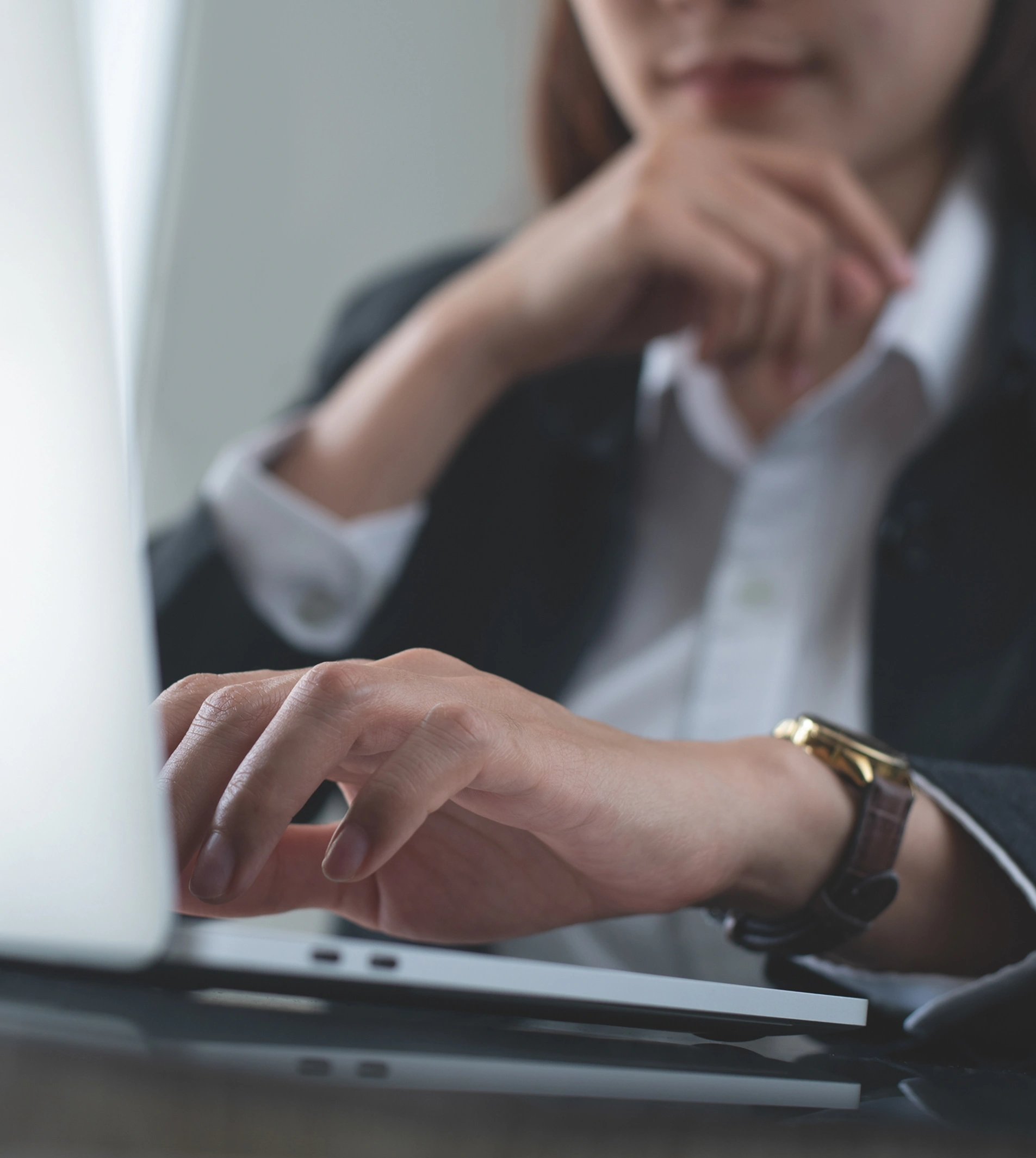 Close-up of a person's hands typing on a laptop, wearing a dark blazer and a gold-faced wristwatch.