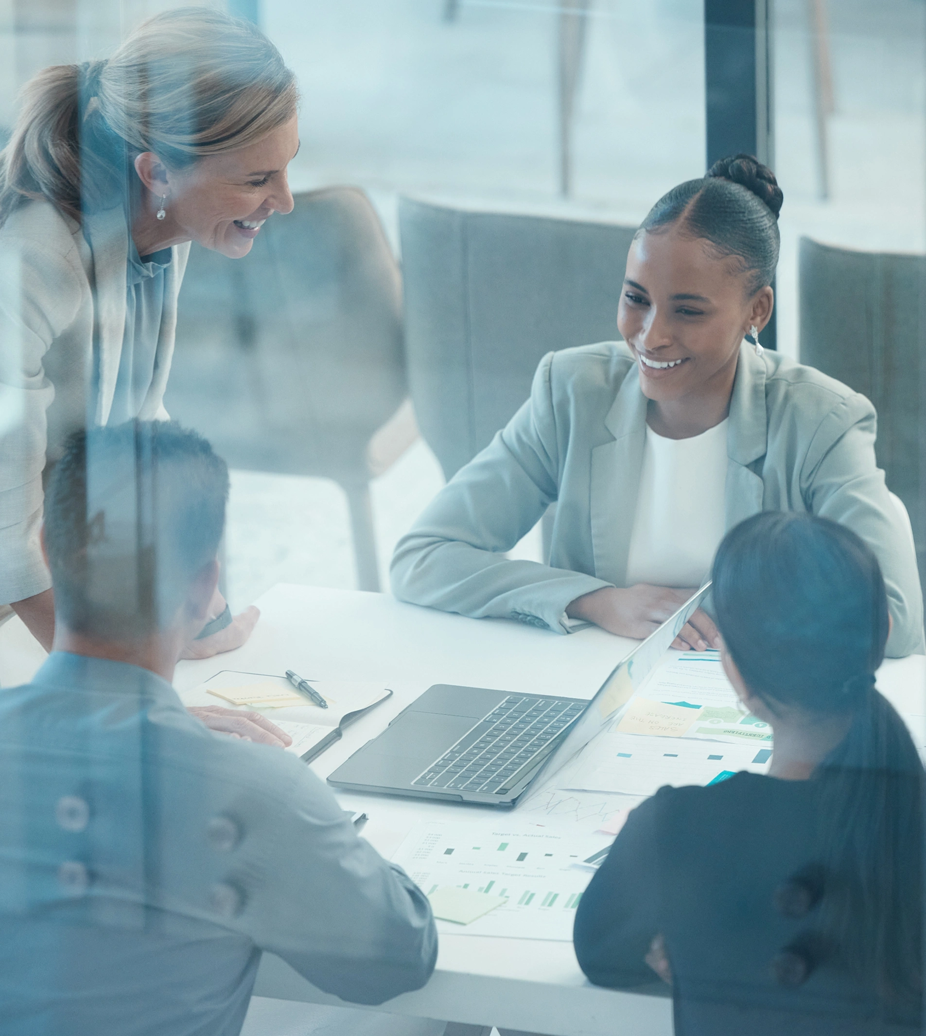 Four professionals in a modern office collaborate around a table with a laptop and reports, engaged in discussion.