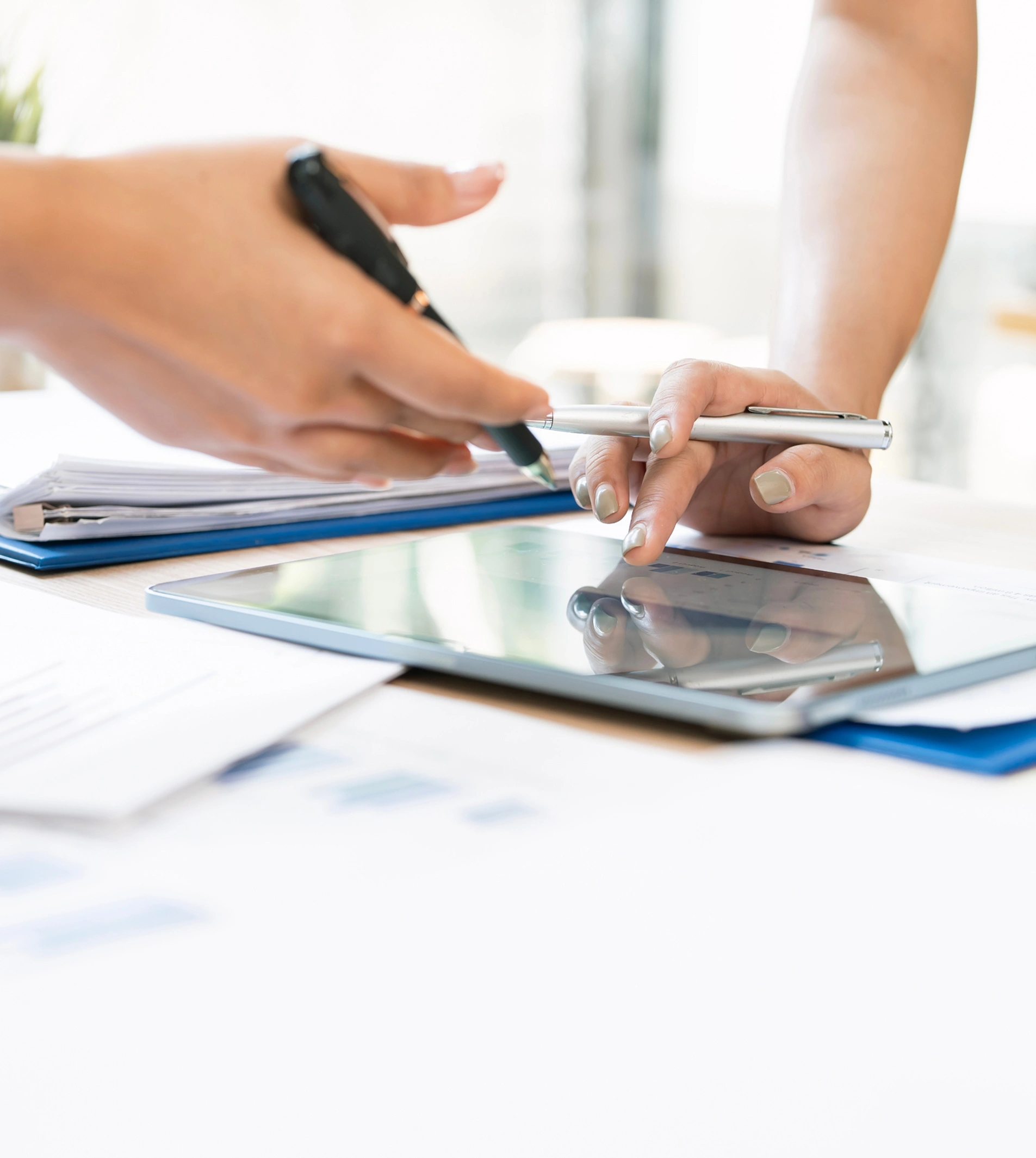 Two hands collaborating over a tablet computer and a stack of documents on a desk.