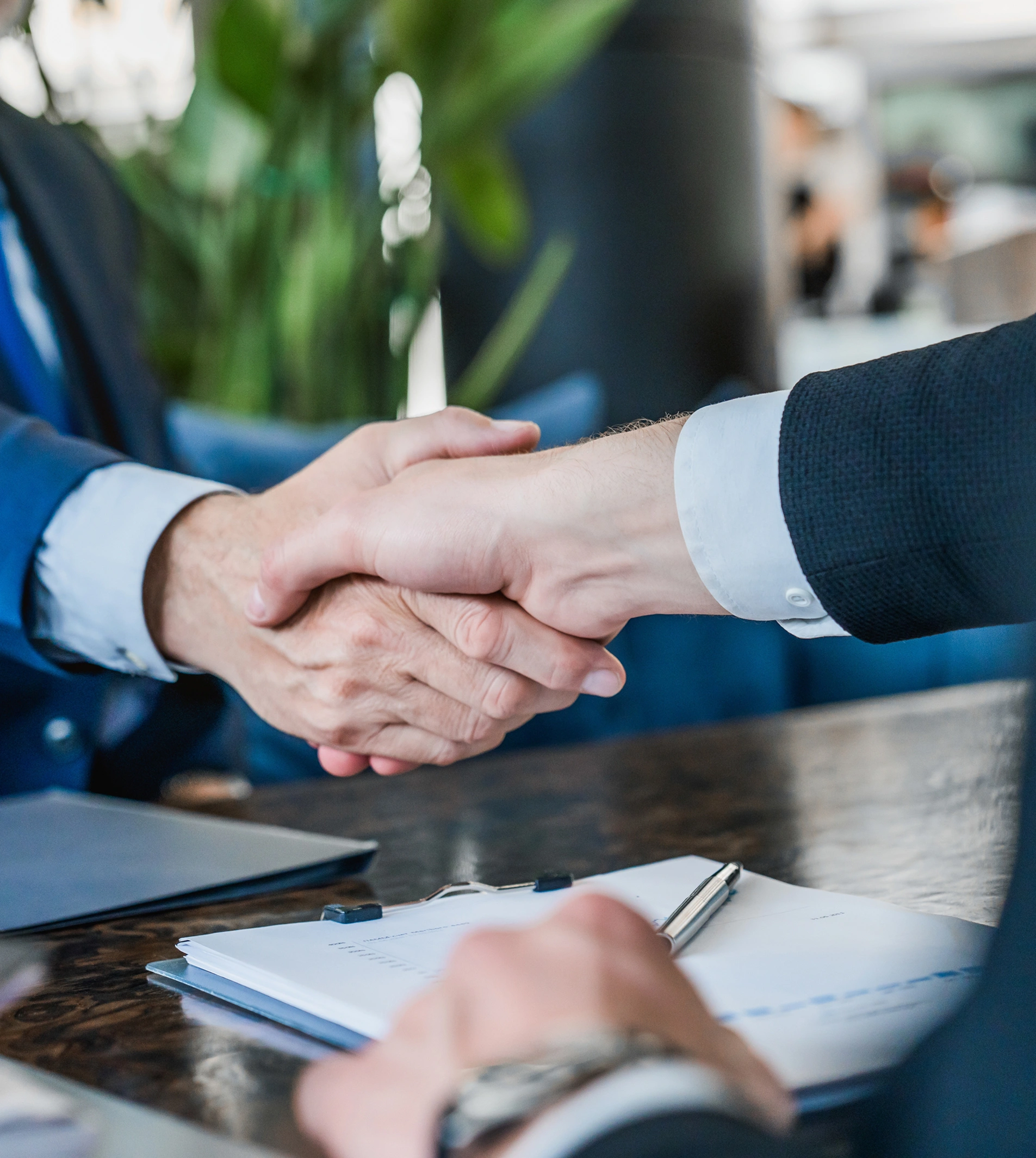 Two individuals in business attire shake hands over a table with documents and a pen.