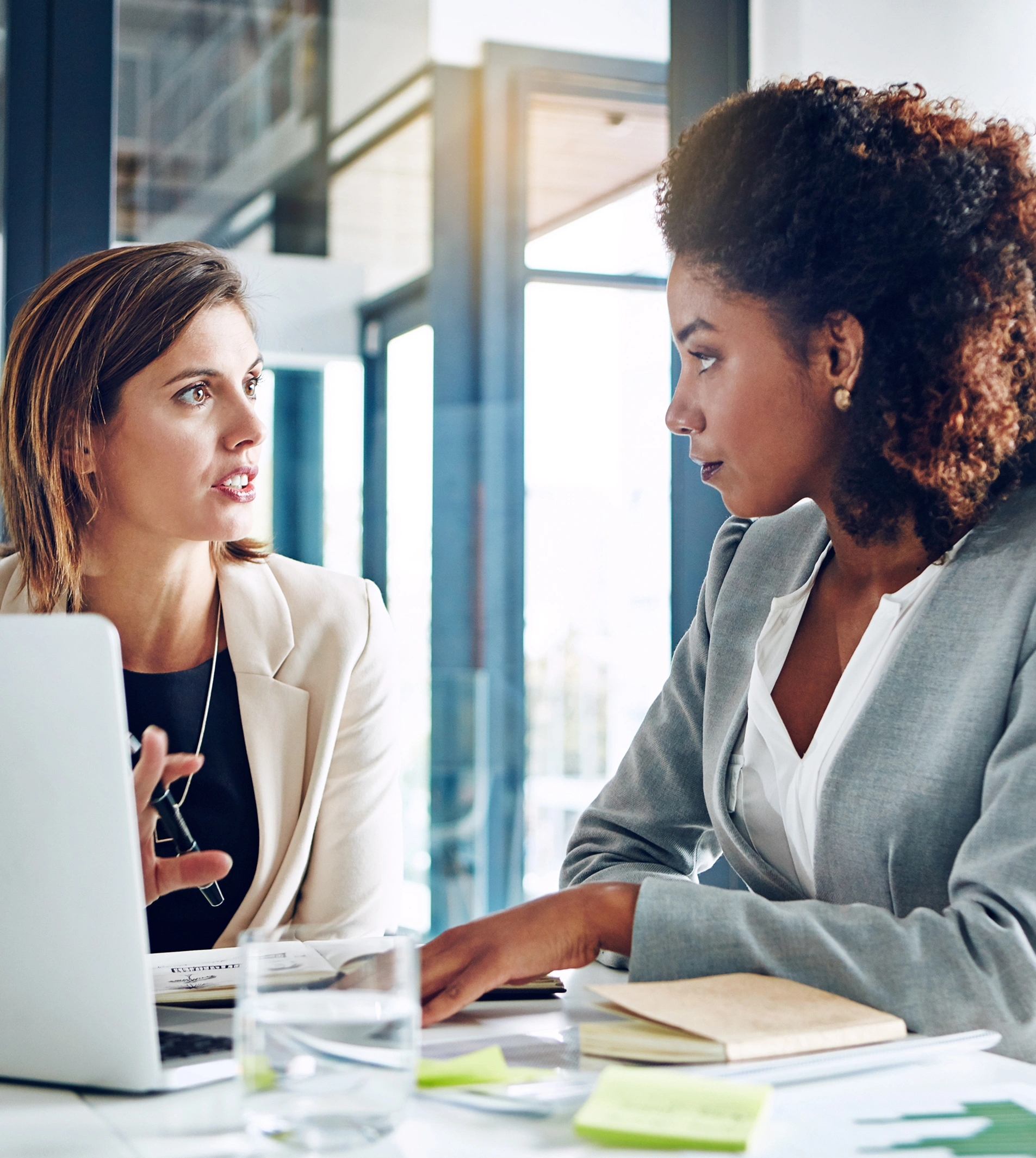 Two women in business attire are engaged in a discussion at a table with laptops and papers in an office setting.